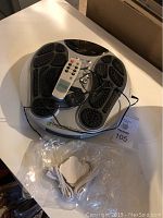 Foot massager with black textured footpads, control panel with buttons, remote control resting on top, and wired adhesive electrode accessory below the unit.