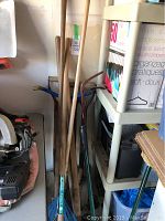 Photo showing garden tools with wooden handles leaning against wall and shelf, including metal rake and plastic leaf rake handles.