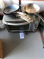 Oster convection oven with three stainless steel pans on top: two frying pans with long handles and one round pan.