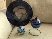 Photo showing three hand blown glass pieces: a large dark blue round hanging disc, a multicolored Murano glass paperweight, and an iridescent hanging ball on a stand
