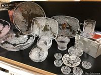 Crystal serving pieces arranged on shelf, featuring etched rose floral designs in plates, bowls, mugs, and vase with original boxes in background.