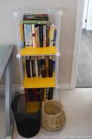 White metal wire shelving with two yellow shelves filled with books placed against a wall next to a grey railing. Two wastebaskets are beside the shelving unit: a black wire mesh and a wicker basket.