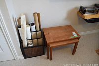 Two stacked solid teak nesting tables placed near a black holder with rolled papers, showing wear and a green label on one table.