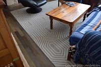 Photo showing the full view of the grey and white geometric patterned area rug under a wooden coffee table and adjacent to black furniture.