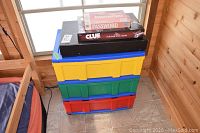 Stacking storage bins in yellow, green, and red, bottom bin taped and damaged, with game boxes on top