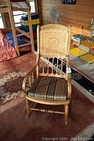 Front view of the antique solid wood rocking chair with patterned cushion on seat, set on a brown carpet in a wood-paneled room.