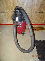 Red 13 gallon Shop Vac unit with flexible hose and power cord attached, positioned on floor against insulation wall.