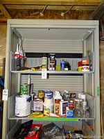 Shelving unit with six unopened tubes of caulking, various containers of paint, primer, stain, wood filler, Danish oil, varsol, lubricating oil, and assorted tape rolls mostly open.