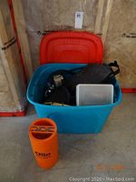 Wide view of blue bin containing various diving equipment including fins, regulator, and wetsuits plus the orange Olin marine signal kit on the floor next to it.