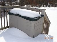 Rubbermaid outdoor deck box covered in snow, beige body with dark green lid, angled view showing contour and hinge design
