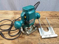 Makita plunge base router with power cord and removable fence on wooden surface.