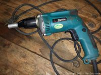 Full view of Makita drywall screwdriver on wooden surface showing teal body, black chuck, and electrical cord coiled around.