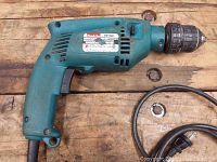 Side view of blue-green Makita HP1501 hammer drill laid on wood surface, showing ergonomic handle, trigger, and cord.