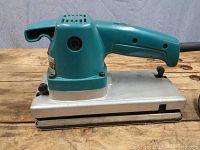 Side view of teal Makita finishing sander with aluminum base and power cord on wood surface