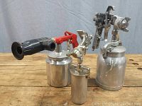 Five compressed air spray guns arranged on wooden surface showing metal containers, triggers, and nozzles, including a red handled sand blasting gun with media inside container.