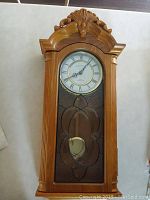 Front view of solid wood wall clock with carved top and leaded beveled glass front showing pendulum and clock face with Roman numerals.