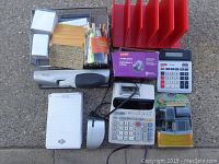Photo of various small office items including stapler, hole punches, calculators, desk organizer with pens and sticky notes, mouse and notepad.