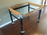 Full view of metal work bench frame from an angle, showing the wooden top frame and metal legs with casters.