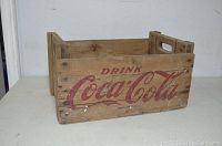 Front side of wooden Coca Cola crate showing red 'Drink Coca Cola' script text with wear and paint chipping.