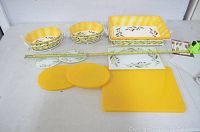 Ceramic baking dishes, lids, glass trivets, wire racks, and yellow plastic boards laid out with measuring tape for scale.