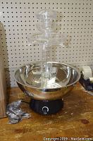 Clear acrylic multi-tiered beverage fountain with large stainless steel bowl and black base with flow control knob, sitting on wooden surface.