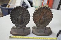 Two cast iron vintage bookends shaped as peacocks with large fanned tails, placed on a white surface showing their size with a measuring tape in front.