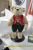Front view of the plush teddy bear standing on a display base, showcasing red vest with gold chains and green pants, with measuring tape showing height around 14.5 inches.