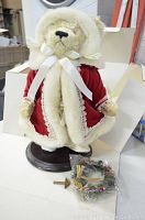 Photo showing the teddy bear in red coat with white fur trim, seated on a display stand, accompanied by a small decorative wreath and an unassembled wooden stand piece in plastic.