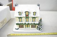 Photo showing the front view of the Village Bakery model building with snow-covered roof, green awnings, lit windows, and a decorated Christmas tree on the side. Includes measuring tape showing size.