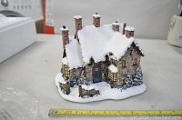 Photo of lighted miniature Christmas village house sculpture from Thomas Kinkade's Village Christmas Collection showing detailed snowy roof, warm lit windows and chimney details, measuring approx 8 inches wide.