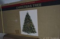 Photo of a large brown cardboard box with an image of a fully assembled pre-lit 7' Canyon Fir Christmas tree on it, showing warm white lights on green branches. Box labeled 'Flame Retardant Christmas Tree'.