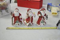 Three Royal Albert Santa Claus porcelain ornaments in red and white attire with gold accents, displayed on a table with a measuring tape for scale, about 5-6 inches tall each.