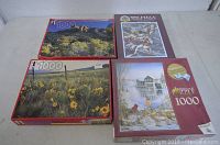 Four unopened 1000-piece jigsaw puzzles with slight damage or soil on outer boxes, various nature and scenic images.