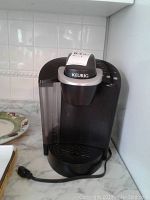 Front view of black Keurig single-serve coffee maker with visible water reservoir, control buttons on top, power cord visible.