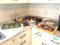 Kitchen countertop showing all items including cast iron skillet with decorative handle, French Press box, serving plates with Christmas and floral designs, butter spreaders, marble trivet, candy dish, salt shaker, and steak knives in tray.