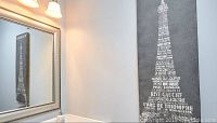 Paris themed bathroom decor on wall including stretched canvas print of Eiffel Tower made of Paris landmark names, positioned next to a lighted bathroom mirror.