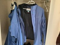 Two blue rain jackets hanging on white hangers inside closet, one Columbia size XL, another Marmot size L