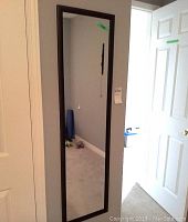 Full view of rectangular wall mirror with dark faux wood frame hung on a wall reflecting part of a room including a floor and a door.