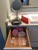Photo of lighted magnifying makeup mirror on bathroom vanity countertop with small gray and red makeup bags. Drawer open showing acrylic vanity organizer and round pink and white bags behind it.