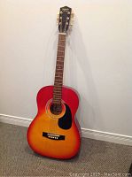 Full view of the sunburst acoustic guitar standing against a wall showing the whole body, neck and headstock.
