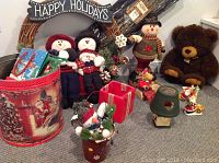 Photo shows the wreath with Happy Holidays banner, two stuffed snowmen, decorated festive bucket, green and red candle holders, and large plush brown bear.
