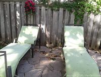 Two metal frame lounge chairs with light green worn cushions placed outdoors on stone patio with wooden fence behind.