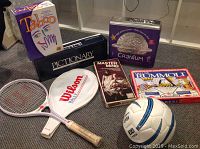 Photo shows all items in the lot arranged on carpeted floor including five board games, soccer ball, tennis racket with cover.