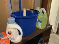 Front view showing large blue mop bucket with mop inserted, Tide detergent jug, and yellow-green Ocean Wet fabric softener jug on brown shelf