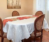 Dining table with white tablecloth, autumn leaf patterned fabric table runner laid down center, surrounded by wooden chairs with upholstered backs and seats.