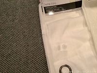 Folded white sheer curtain panels with visible metal grommets and product label showing brand and product type.