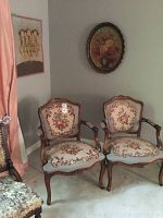 Two needlepoint armchairs with carved wooden frames and floral upholstery placed side by side. Oval framed floral print and rectangular floral painting visible on the wall behind chairs.