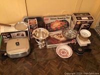 Wide photo showing Oster meat slicer, electric frying pan, roasting pan, pitcher, bowls, coffee maker box, various kitchenware items arranged on floor