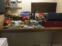 Wide view of a workbench with assorted hardware and tools including toolboxes, flashlights, wrenches, propane torch, vacuum, electrical cords, and packages of switch covers.