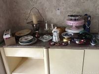 Wide view showing assortment of candle holders, ceramic plates, and figurines on table against wallpaper.